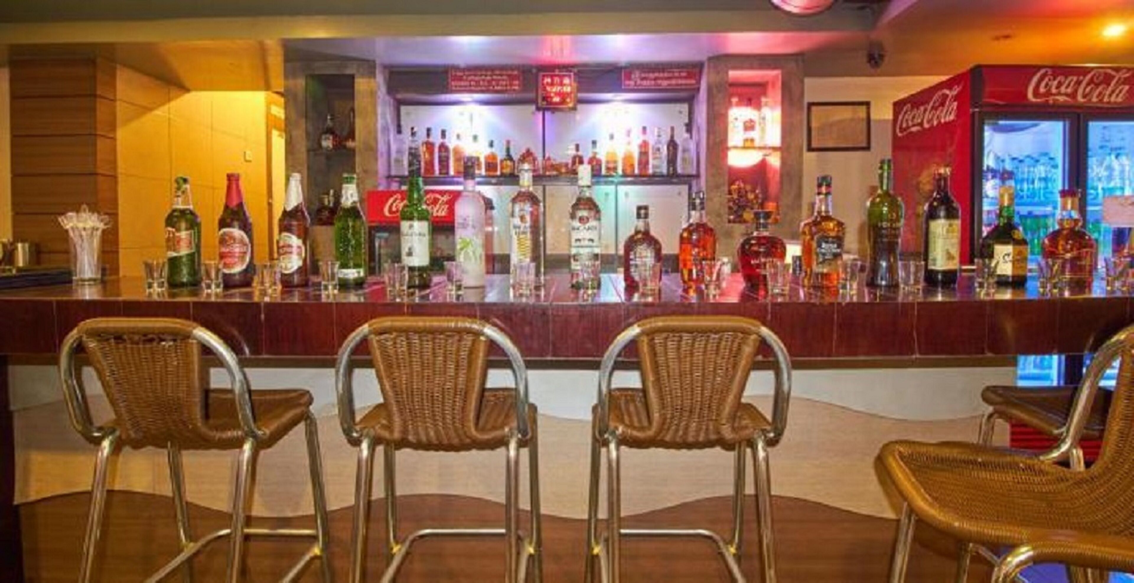 A bar area with multiple liquor bottles, a Coca-Cola refrigerator, and wicker chairs.