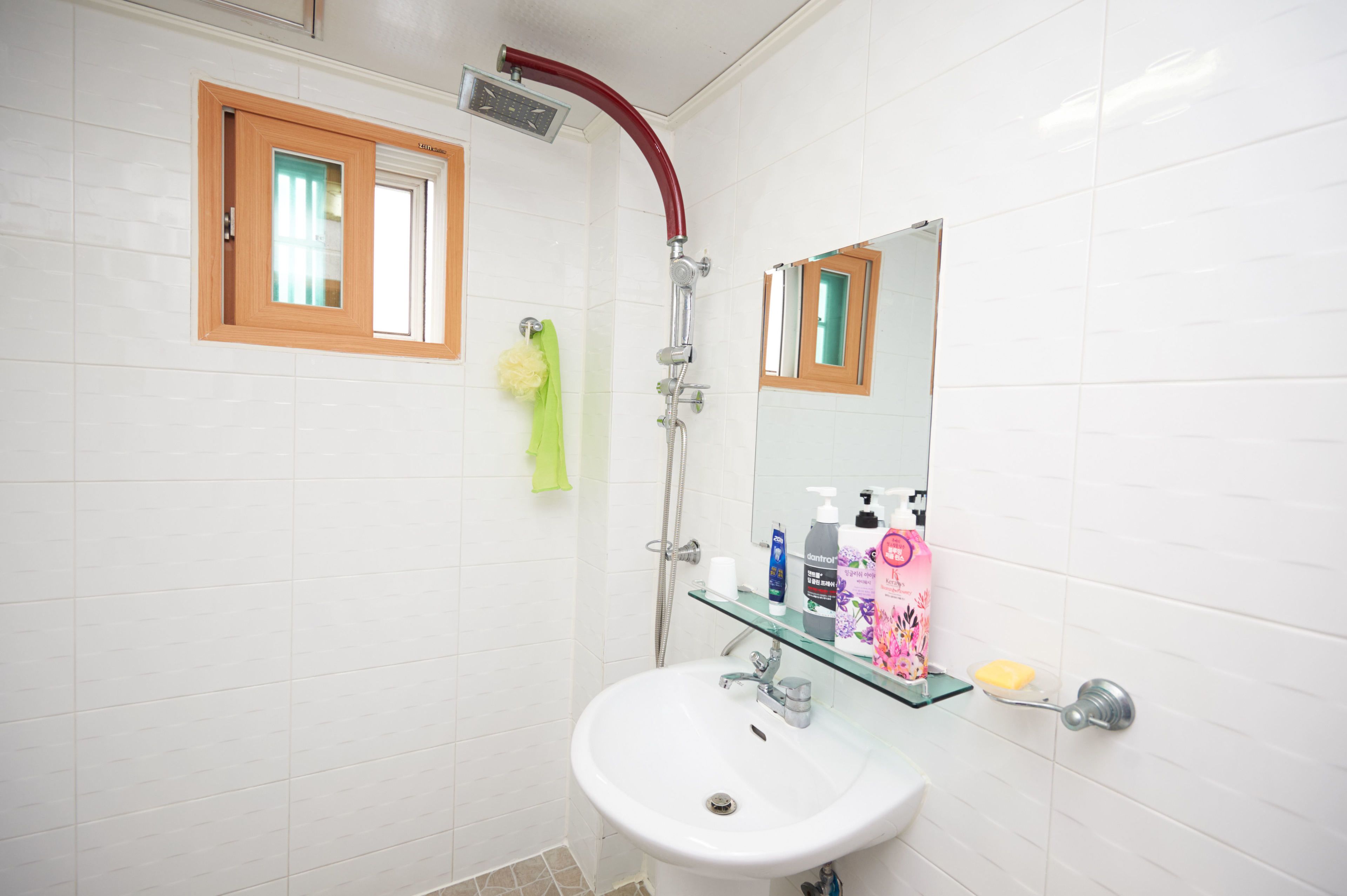 Ein Badezimmer mit einem weißen Waschbecken, einer Glasablage mit Toilettenartikeln, einer roten Duschbrause und einem Spiegel mit Holzrahmen.