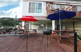 A wooden deck with picnic tables and barbecue grills, a covered area with a blue umbrella, and a building with a balcony.