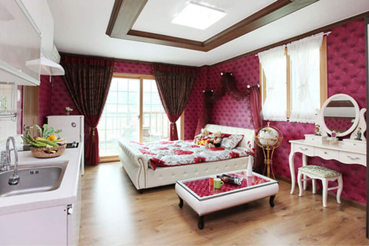 A bedroom with a bed, a vanity with a mirror, a dresser, a small table, and a window with curtains.