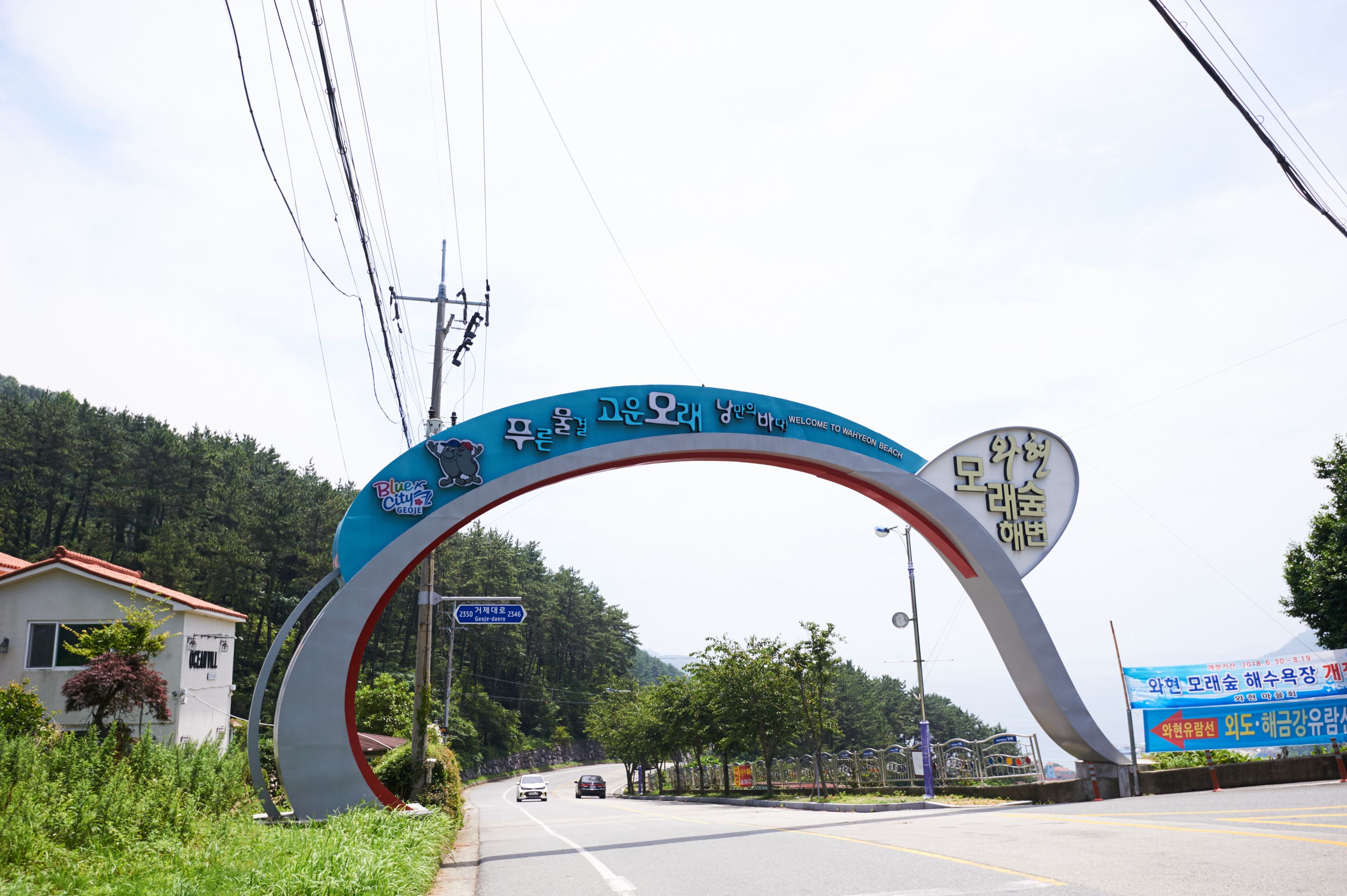 A large archway with foreign text and a clock, leading to a road with cars.