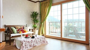 A room with a wooden floor, a sofa with cushions, a small table with a floral tablecloth, a potted plant, and a view through large windows. - Sand Beach Pension (Geoje)