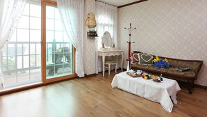 A room with a wooden floor, a white sofa, a small table with a white tablecloth, a chair, a desk with a mirror, and a window with curtains. - Sand Beach Pension (Geoje)