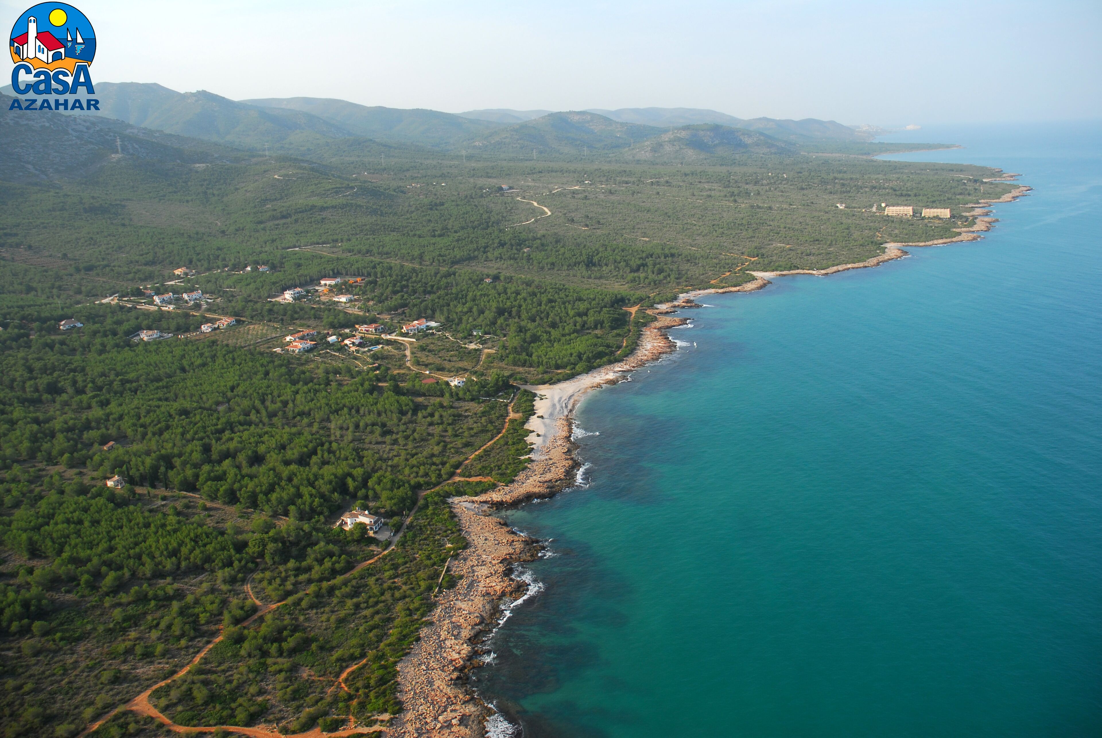 An aerial view of a coastal area with dense forest, a rocky shoreline, and a clear blue sea.