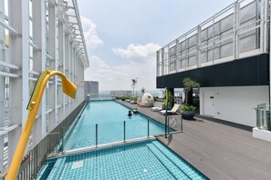 Una piscina en la azotea con agua cristalina y azul, rodeada por una terraza de madera y edificios modernos.