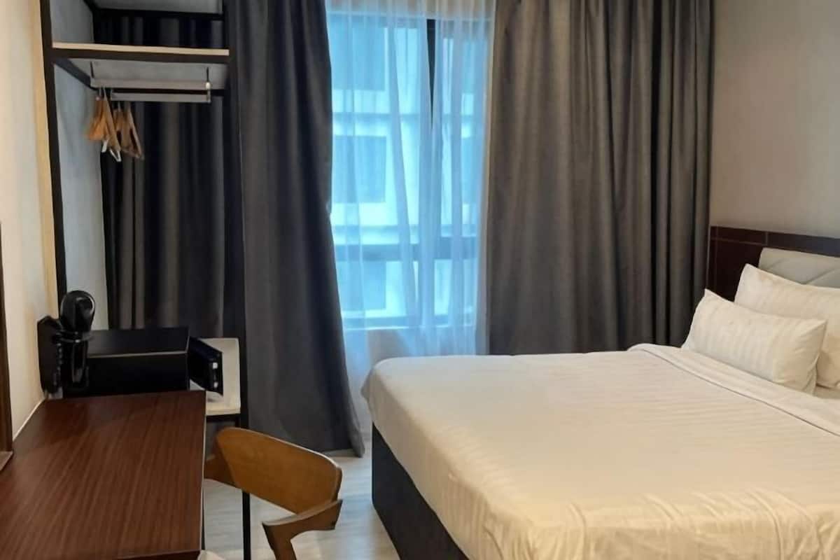 A hotel room with a bed, a desk, a chair, a coat rack, and a window with curtains.