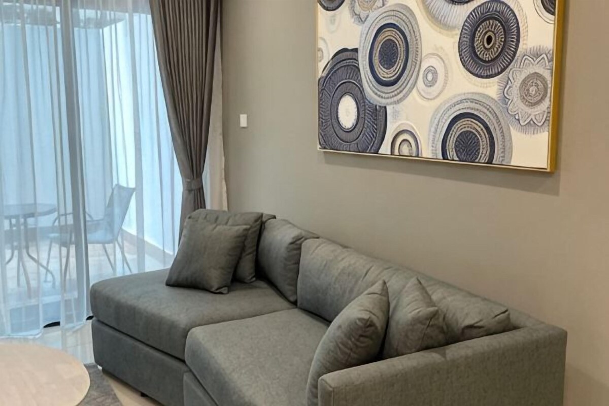 Um sofá cinzento com almofadas, uma obra de arte com padrão na parede e uma porta de vidro que dá para uma área externa.
