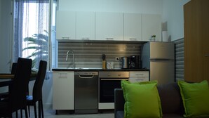 A modern kitchen with white cabinets, a dishwasher, an oven, and a refrigerator. There is a dining area with a table and chairs, and a sofa with green cushions. - Optimist Makarska Town Center (Makarska)