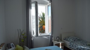 A bedroom with a bed, a nightstand, a window with curtains, and a view of palm trees outside. - Optimist Makarska Town Center (Makarska)