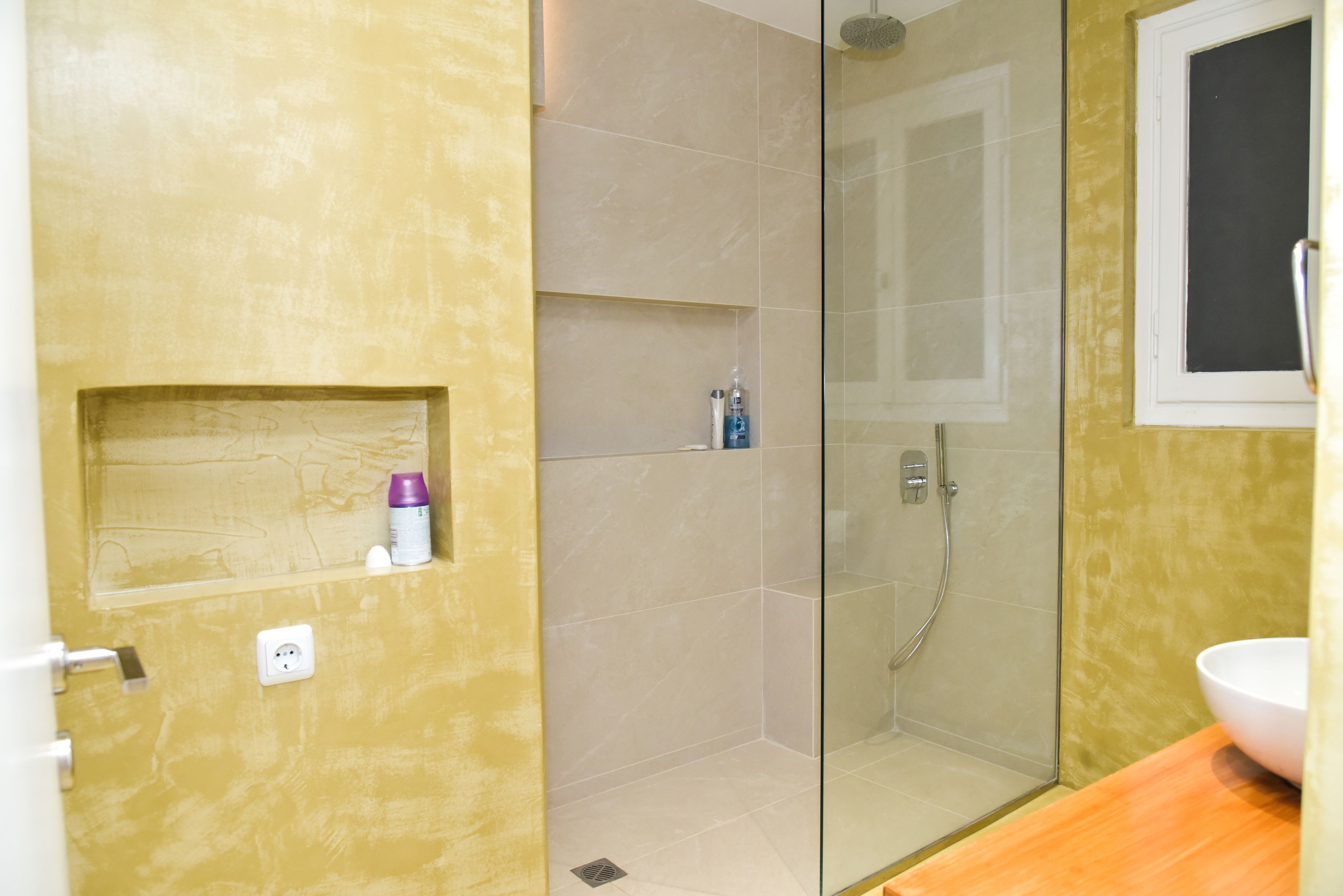 Une salle de bain avec une douche à parois de verre, un lavabo blanc et un mur jaune agrémenté d’une petite étagère.