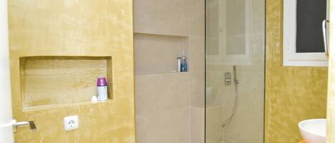 Une salle de bain avec une douche à parois de verre, un lavabo blanc et un mur jaune agrémenté d’une petite étagère.