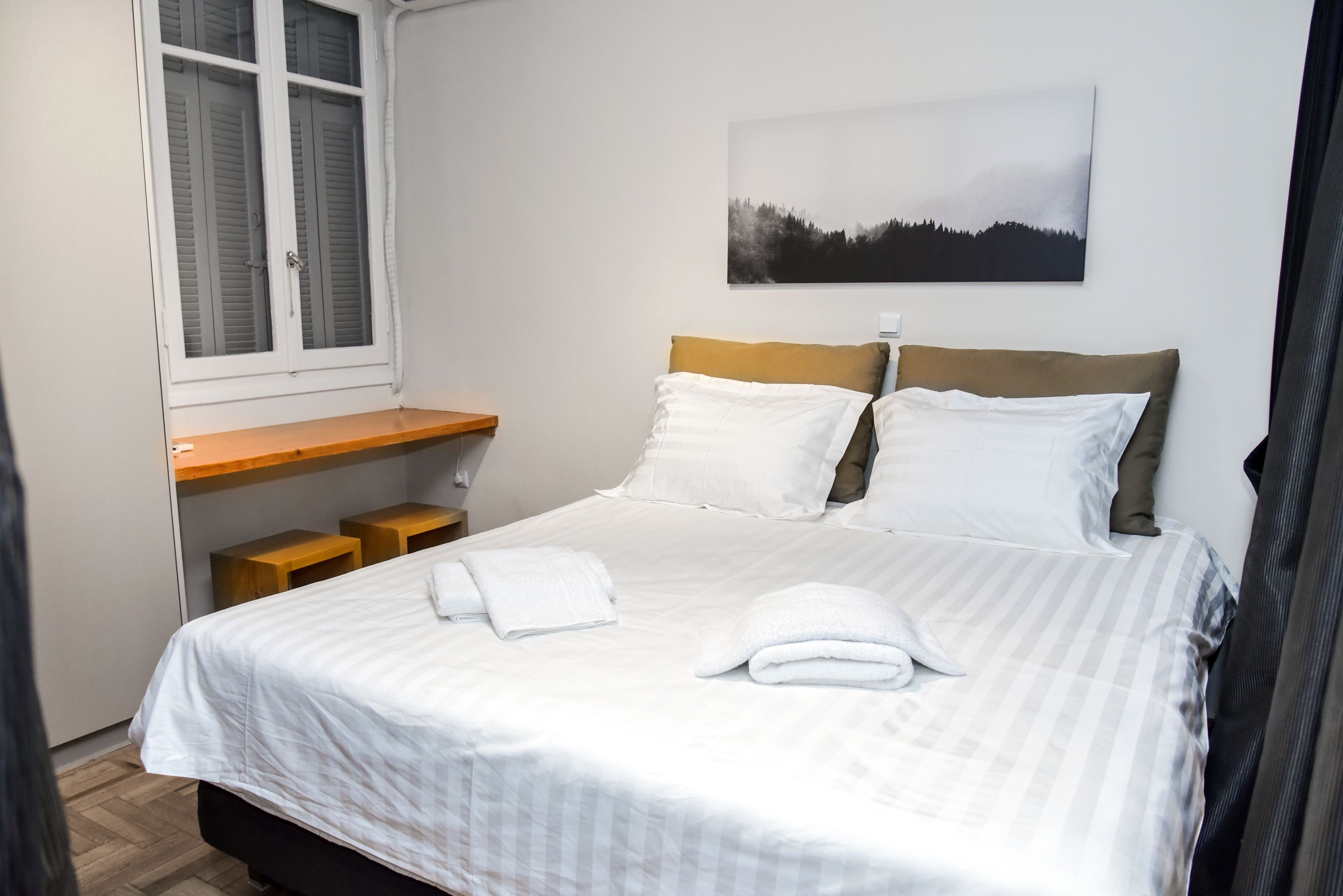 Ein ordentlich bezogenes Bett mit weißen Leinen und Kissen, ein Nachttisch aus Holz, ein Fenster mit Fensterläden und ein Gemälde von einem Wald an der Wand.