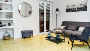 A living room with a grey sofa, a black coffee table, and a grey bench. There is a white shelf with decorative items and a round mirror.