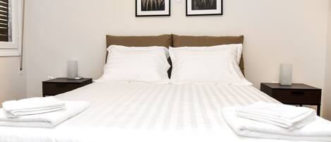 A neatly made bed with white linens and two pillows, flanked by nightstands with lamps, in a room with a plain wall and two framed pictures above the bed.
