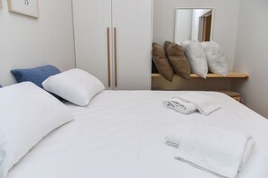 A neatly made bed with white bedding, a pair of folded towels, and a wooden shelf with pillows. - Modern  Apartment at Exarchia 1 bed 2 pers  (Athens)