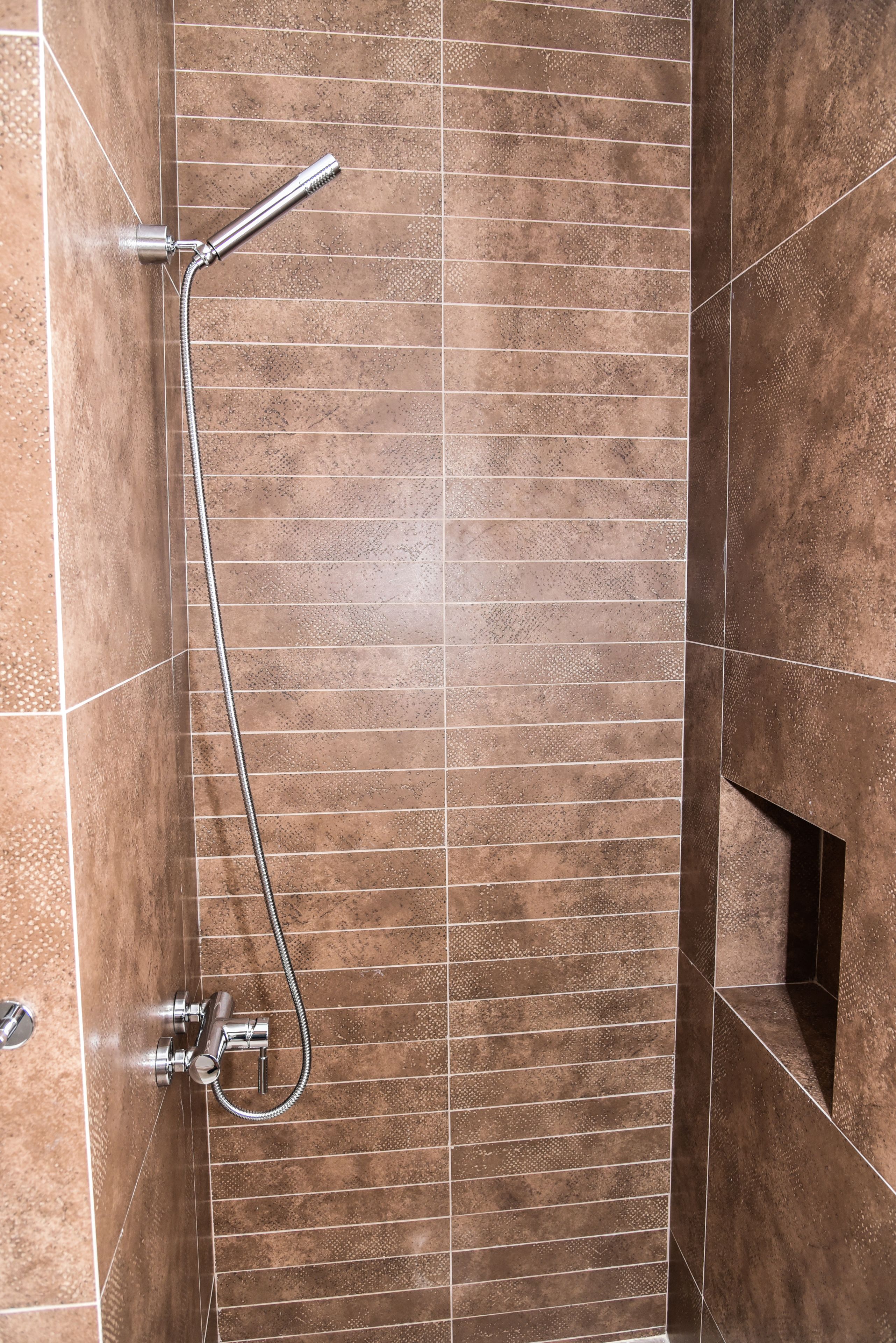 A shower with a handheld showerhead and a wall-mounted faucet, set in a tiled bathroom with a modern design.