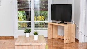 A room with a wooden floor, a TV on a wooden stand, a small table with two potted plants, and a window with blinds. - Modern  Apartment at Exarchia 1 bed 2 pers  (Athens)