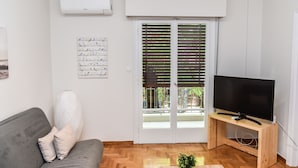 A living room with a grey sofa, a wooden TV stand, a flat-screen TV, a white air conditioner, a glass door with blinds, and a wall-mounted artwork. - Modern  Apartment at Exarchia 1 bed 2 pers  (Athens)