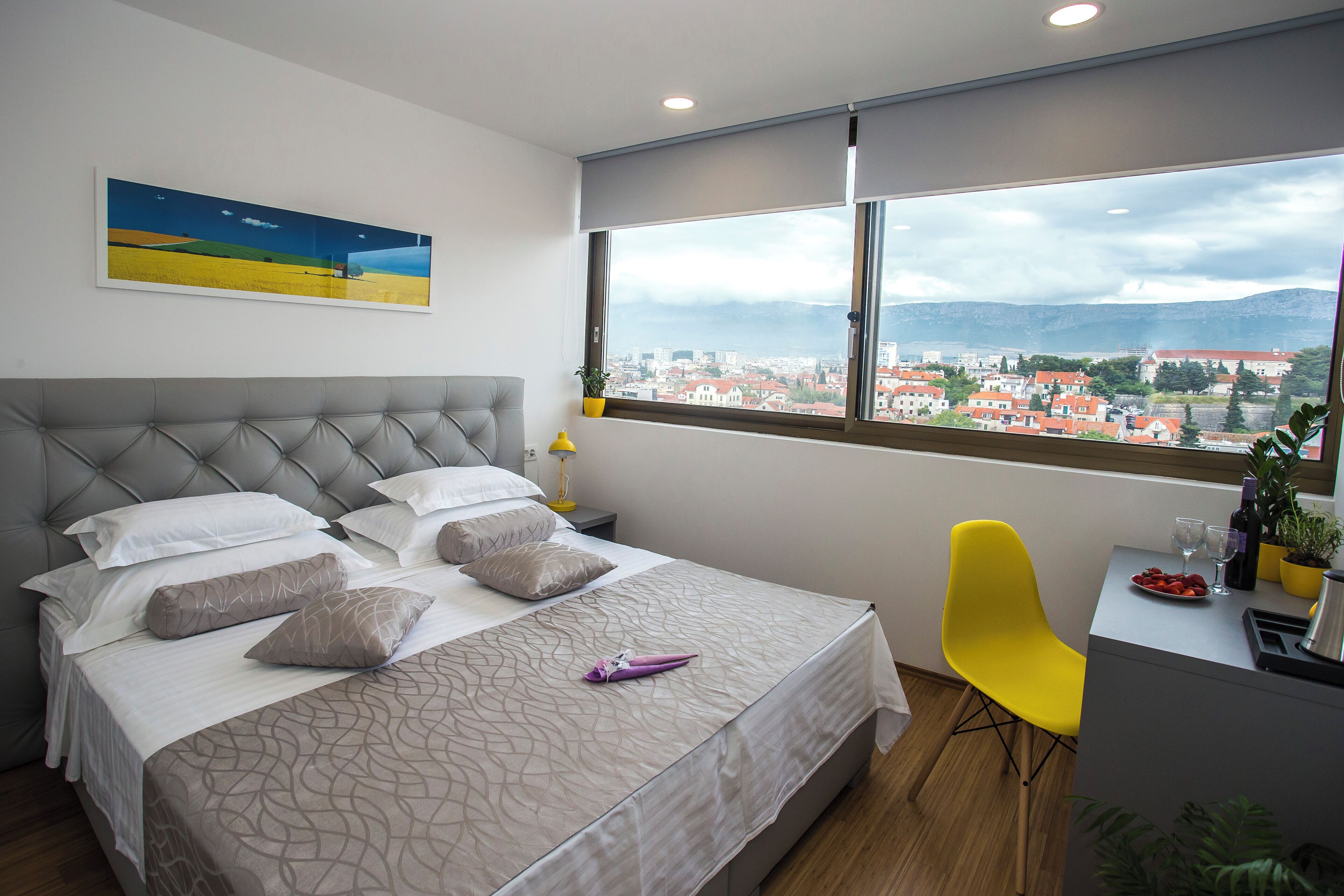 A bedroom with a bed, a desk with a chair, a window with a city view, and a painting on the wall.