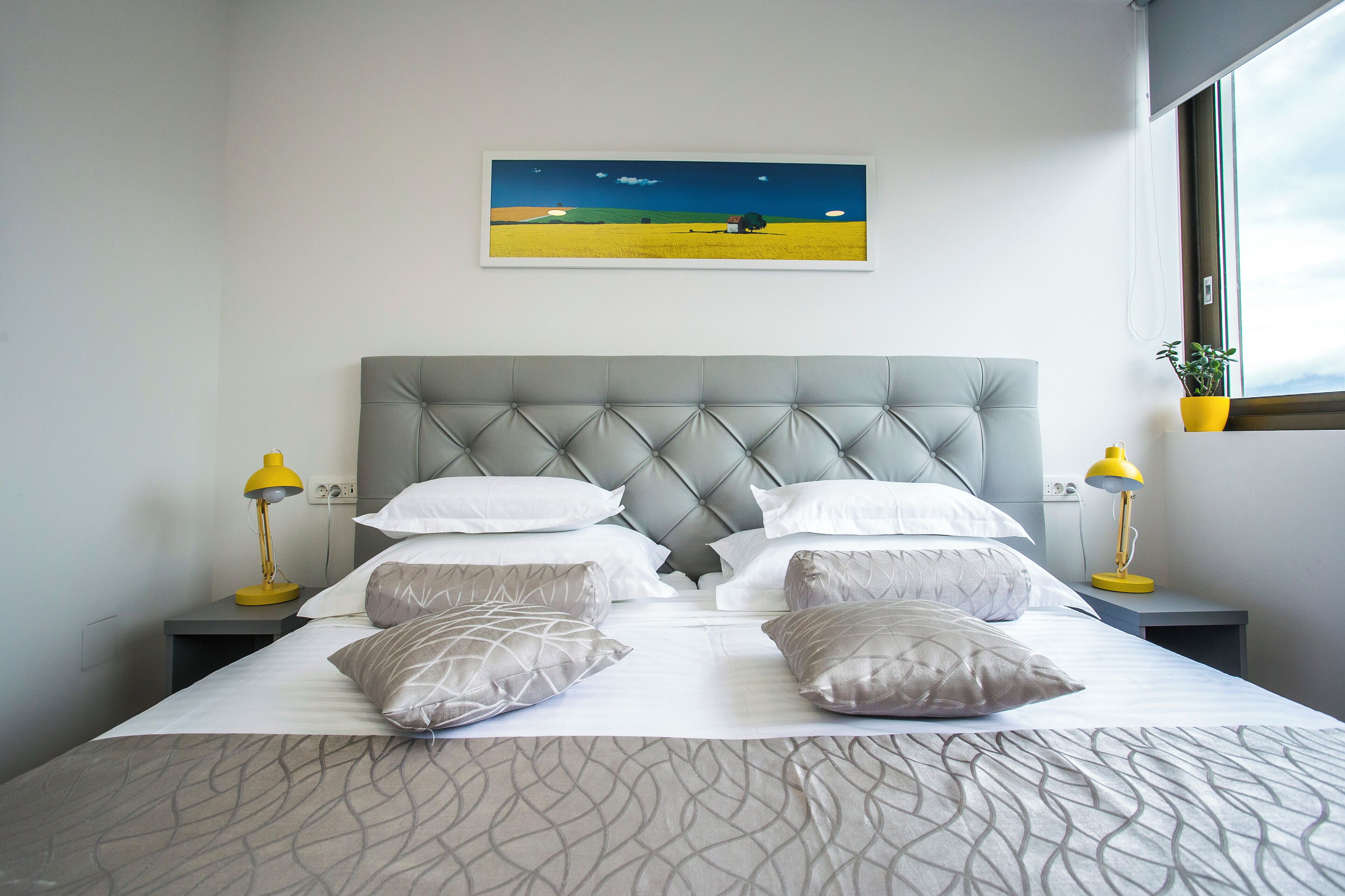 A neatly made bed with a tufted headboard, two bedside tables with lamps, and a painting of a landscape above the bed.