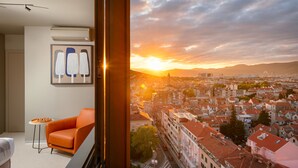 Una habitación con vista a un paisaje urbano al atardecer, una silla moderna color naranja y una mesita con un tazón de aperitivos.