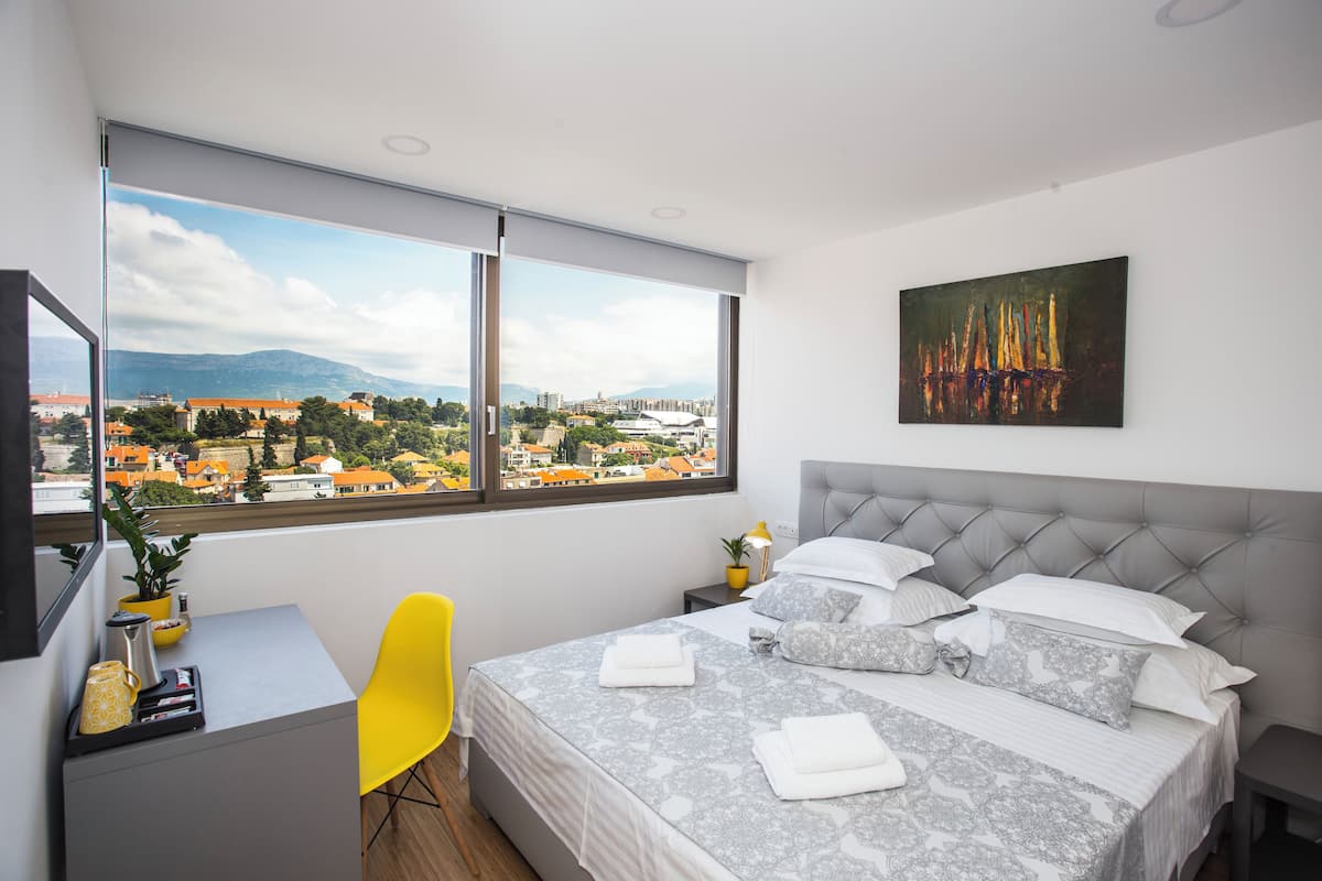A modern hotel room with a large bed, a desk, a chair, and a view of the cityscape through large windows.