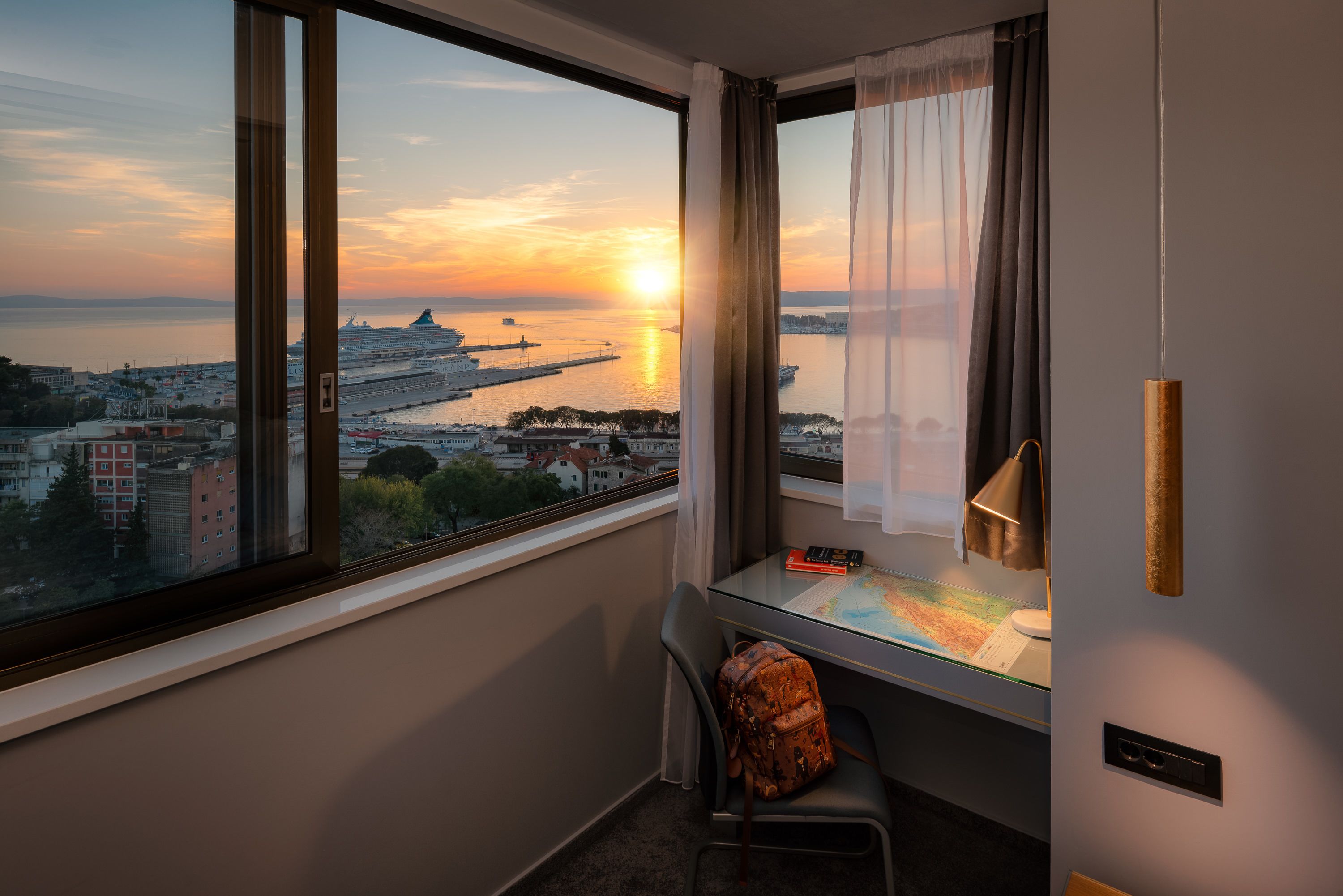 A room with a large window offering a view of a cityscape and a sunset over a body of water.