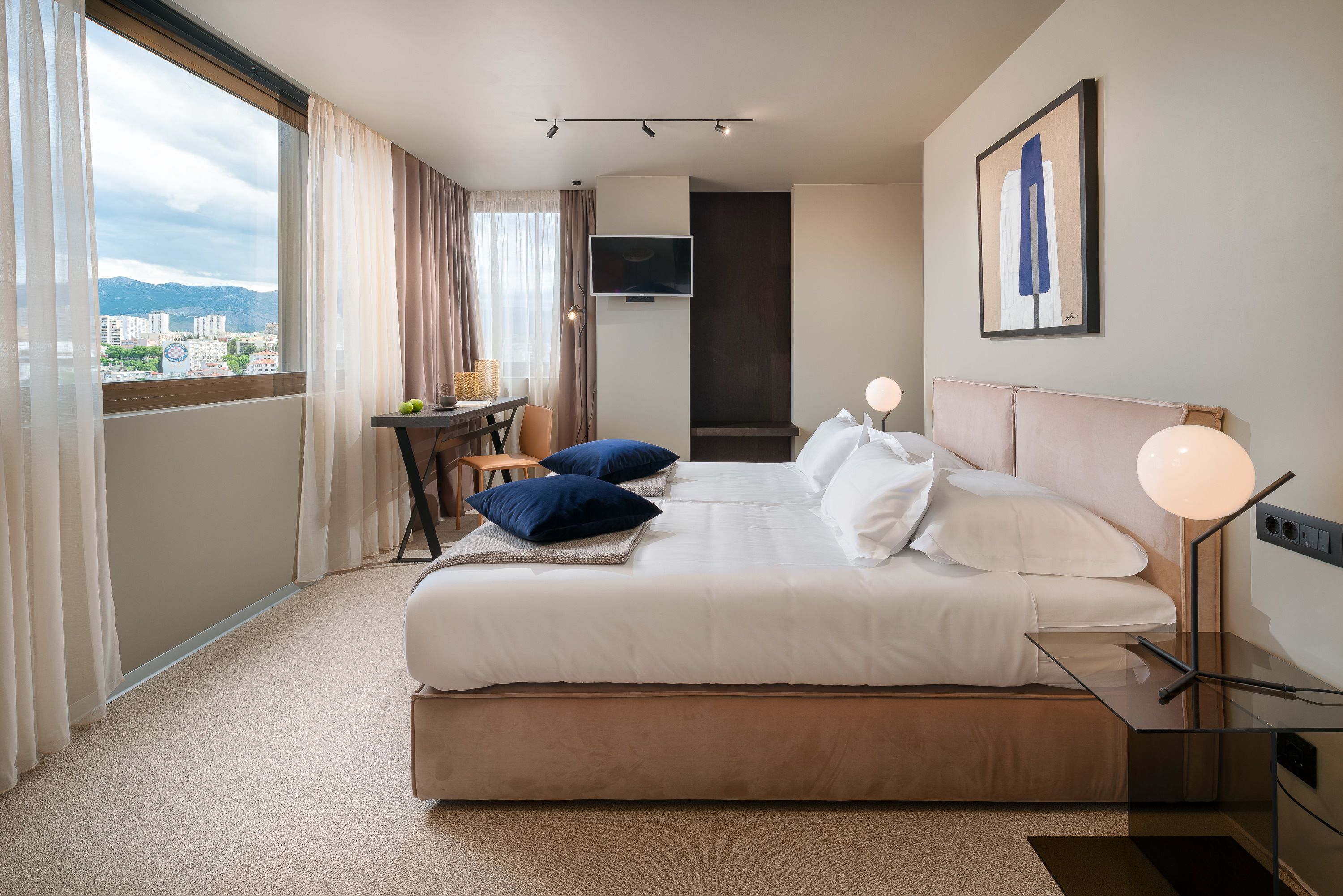 Quarto de hotel com uma cama grande, uma escrivaninha com cadeira, televisão e vista para a cidade.