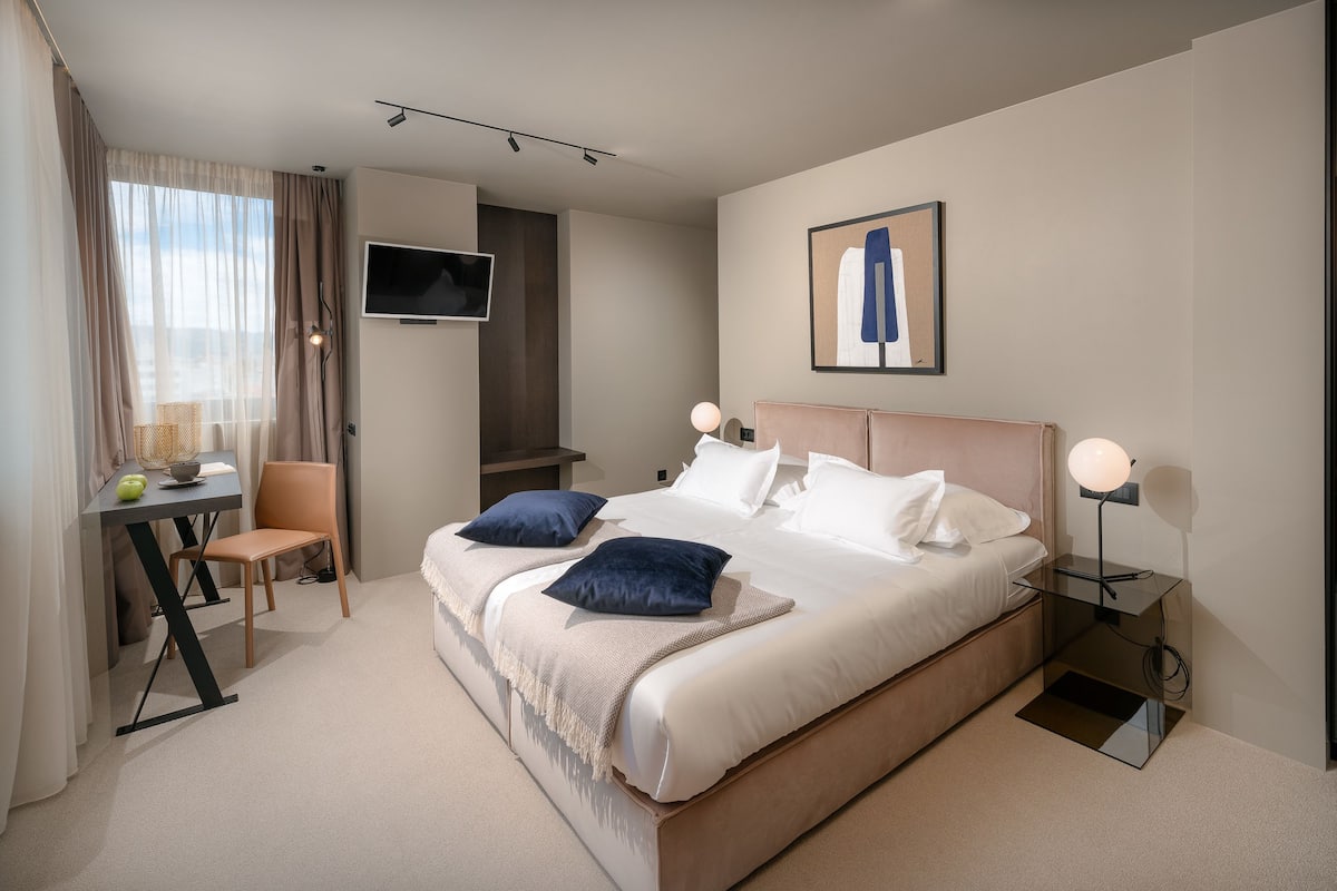 A modern hotel room with a large bed, a desk, a chair, a TV, and a framed picture on the wall.