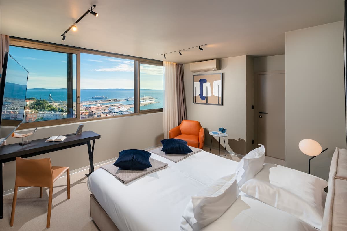 A hotel room with a large window offering a city view, a bed with white linens, an orange chair, a desk with a laptop, and a small table with a lamp.