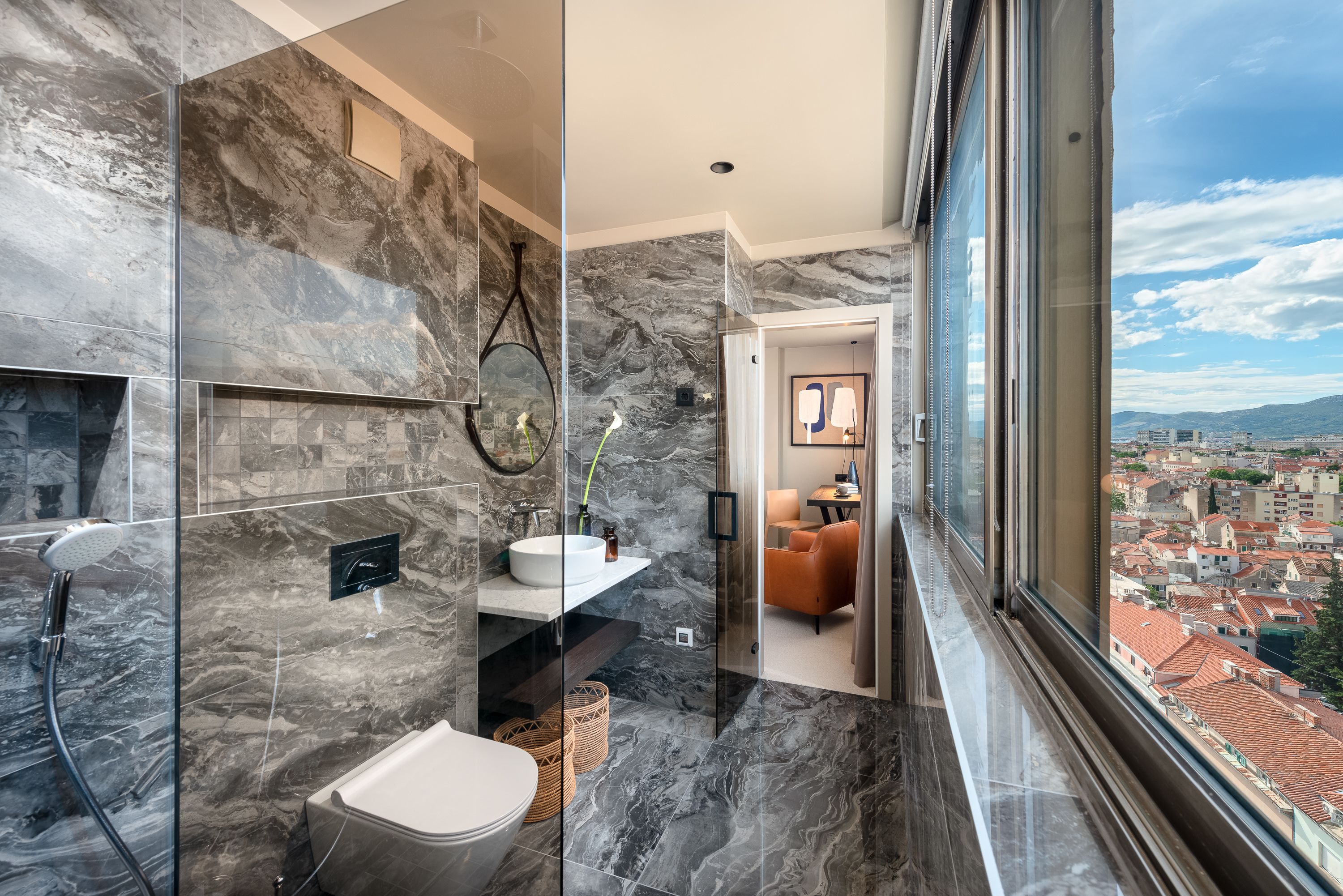A modern bathroom with a marble backsplash, a glass shower enclosure, and a view of the city through a large window.