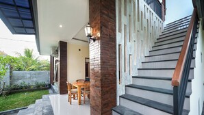 A modern staircase with a wooden handrail, a dining area with a table and chairs, and a brick pillar with a wall-mounted light. - Liam Private Villa (Sukawati)