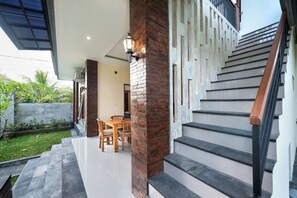 A modern staircase with a wooden handrail, a dining area with a table and chairs, and a brick pillar with a wall-mounted light. - Liam Private Villa (Sukawati)
