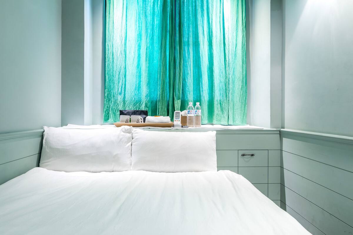 Een net opgemaakt bed in een hotelkamer met een raam, een nachtkastje met toiletartikelen en een bijzettafeltje.