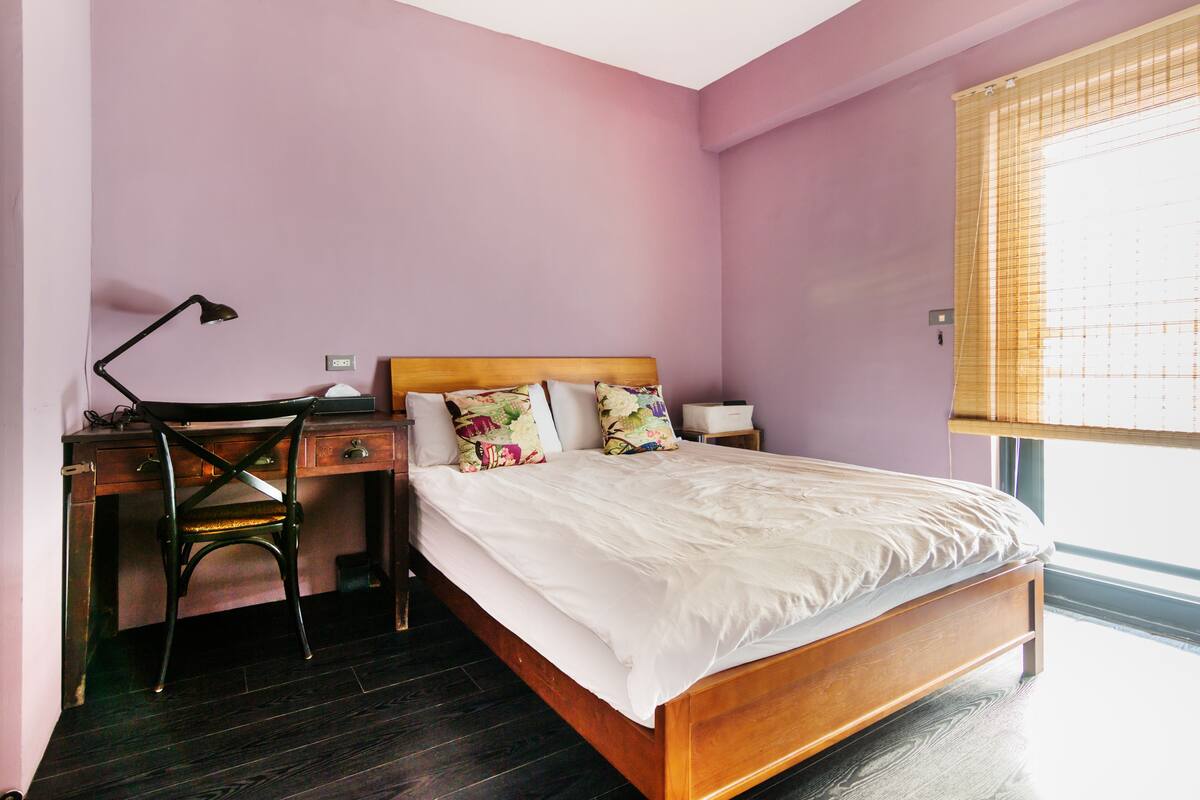 Een slaapkamer met een houten bed, een bureau met stoel en een raam met jaloezieën.