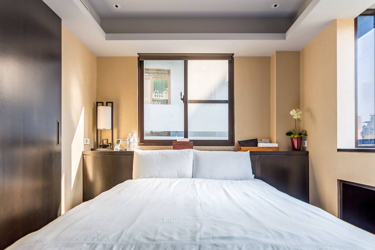 Een hotelkamer met een groot bed, een nachtkastje met een lamp, een raam met uitzicht op gebouwen en een kleine plant op het nachtkastje.