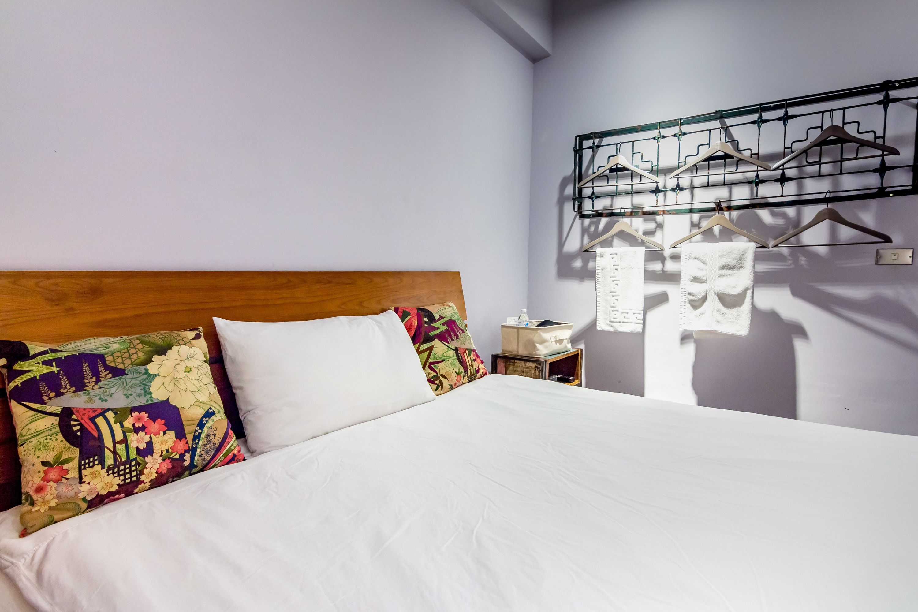 Una cama bien hecha con sábanas blancas, cabecera de madera y almohadas de colores. Se ve un perchero con perchas a la derecha.