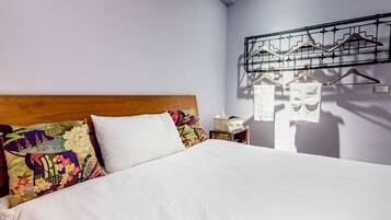 Una cama bien hecha con sábanas blancas, cabecera de madera y almohadas de colores. Se ve un perchero con perchas a la derecha.