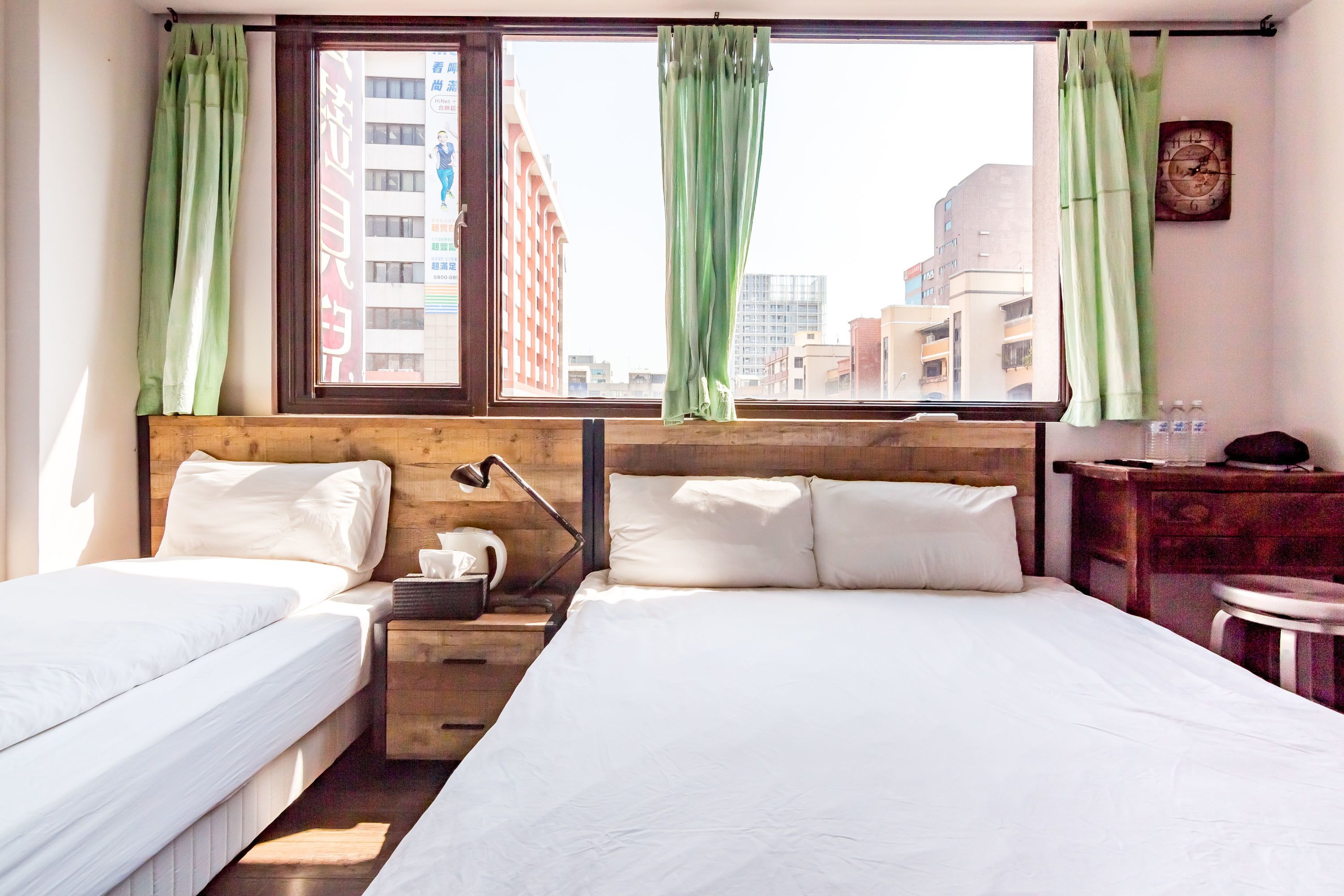 A hotel room with two beds, a wooden headboard, a nightstand, a lamp, and a view of buildings through the window.