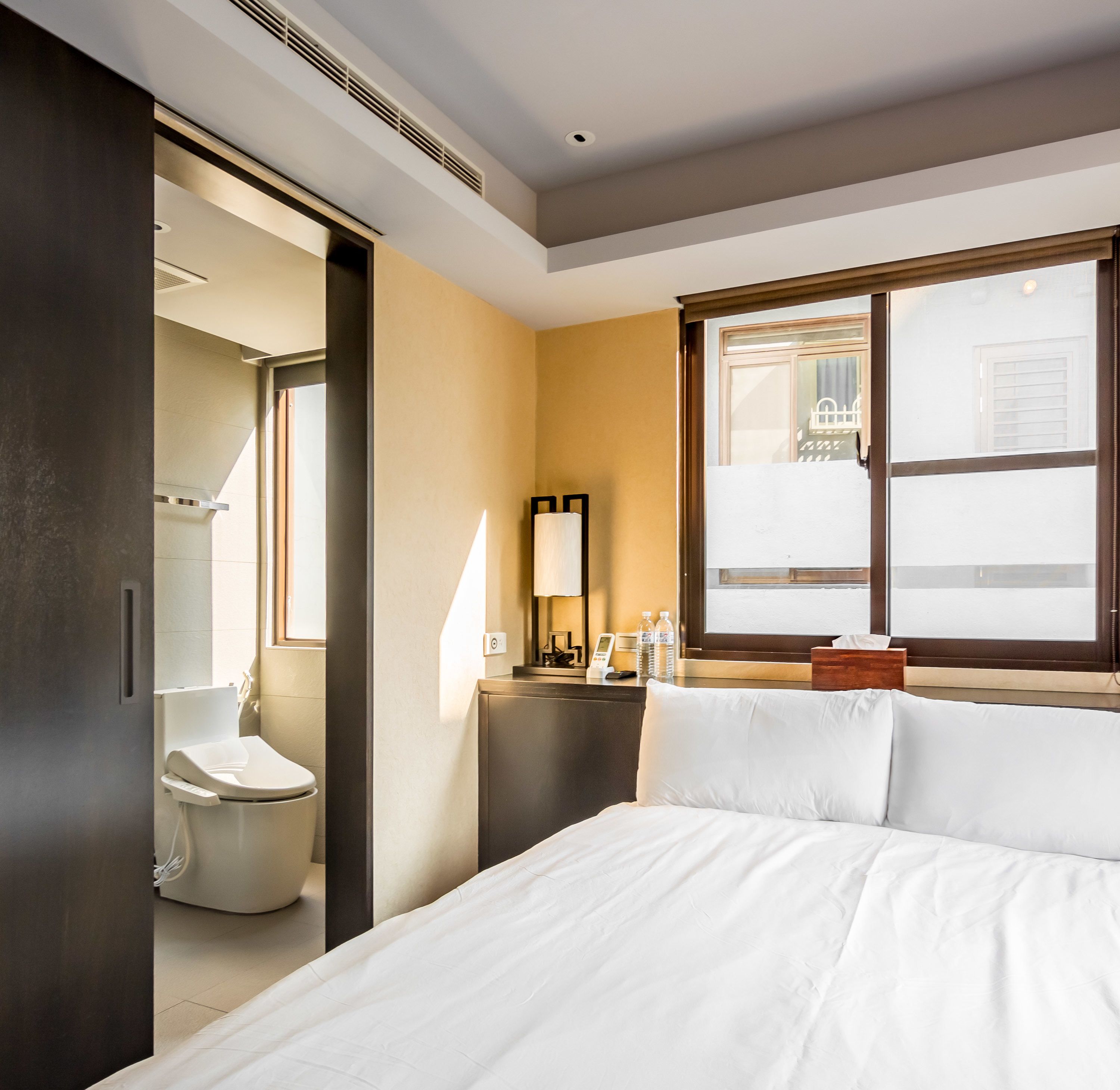A hotel room with a bed, a bedside table with a lamp, a window with a view, and a bathroom with a toilet and sink.