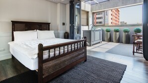 Quarto com cama de madeira, banheiro com pia e plantas em vasos, e varanda com vista para edifícios.