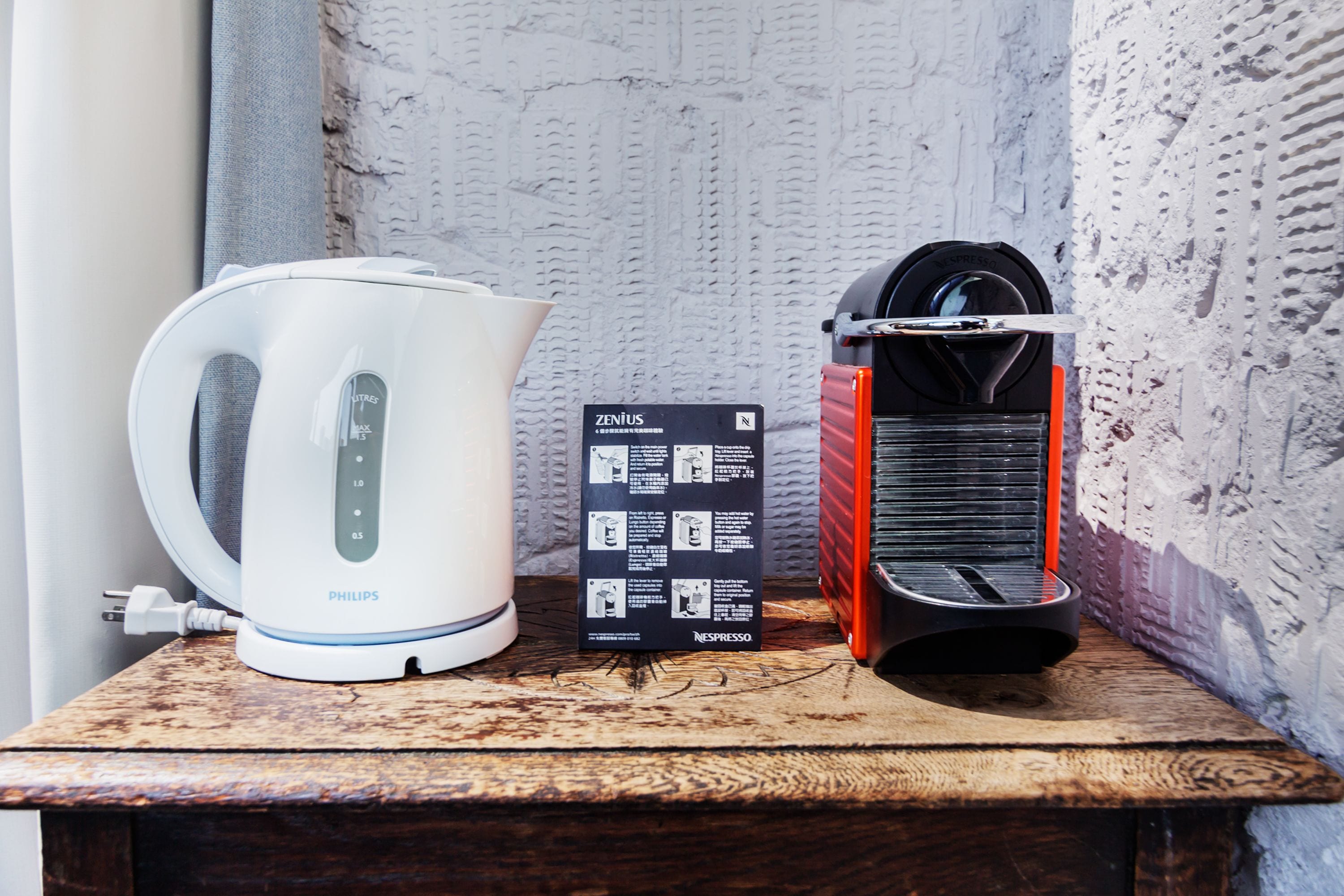 A white Philips kettle, a black and red Nespresso machine, and a user manual on a wooden surface against a textured wall.