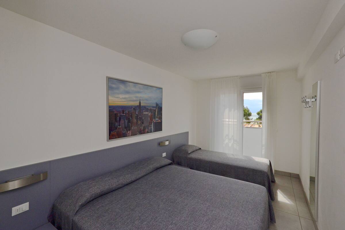 A hotel room with two beds, a cityscape painting, a window with curtains, and a view of the outdoors.