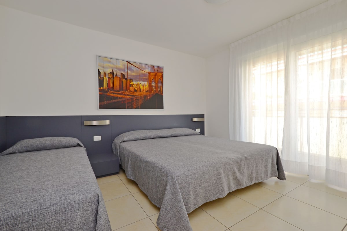 A hotel room with two beds, a cityscape painting on the wall, and sheer curtains.