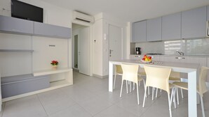 A modern kitchen with white cabinets, a built-in oven, and a dining area with four chairs. - Appartamenti Mare (Lignano Sabbiadoro)