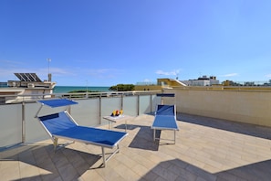 A rooftop terrace with two blue lounge chairs, a small table with a drink, and a view of the sea.