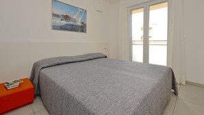 A bedroom with a bed, a painting, a window, and a red side table. - Appartamenti Mare (Lignano Sabbiadoro)