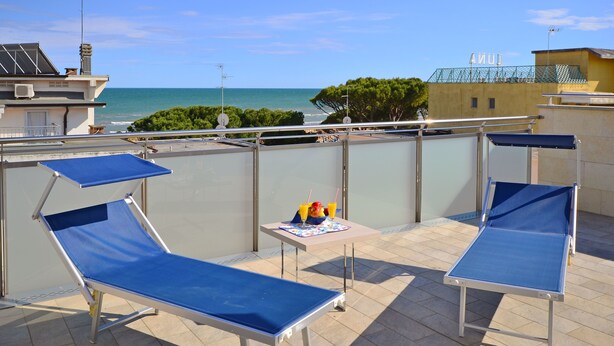 A rooftop terrace with two blue sun loungers, a small table with a drink and fruit, and a view of the sea. - Appartamenti Mare (Lignano Sabbiadoro)
