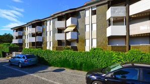A multi-story residential building with balconies, a parking area, and a hedge in front.