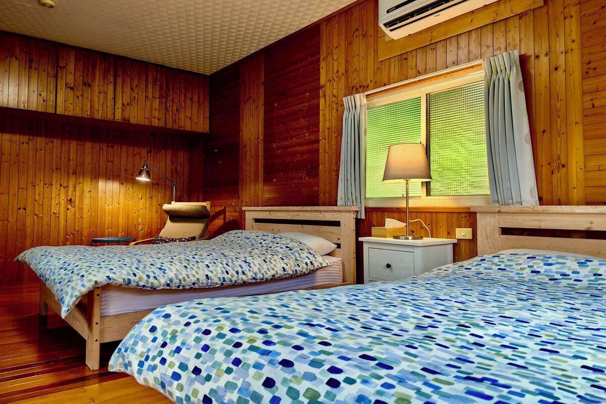 A room with wooden walls, two beds with patterned bedding, a bedside table with a lamp, and an air conditioning unit.